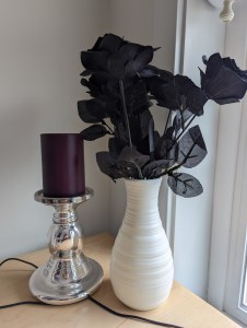 Schwarze künstliche Rosen in einer weißen Vase stehen neben einem silbernen Kerzenständer mit einer lilafarbenen Kerze.