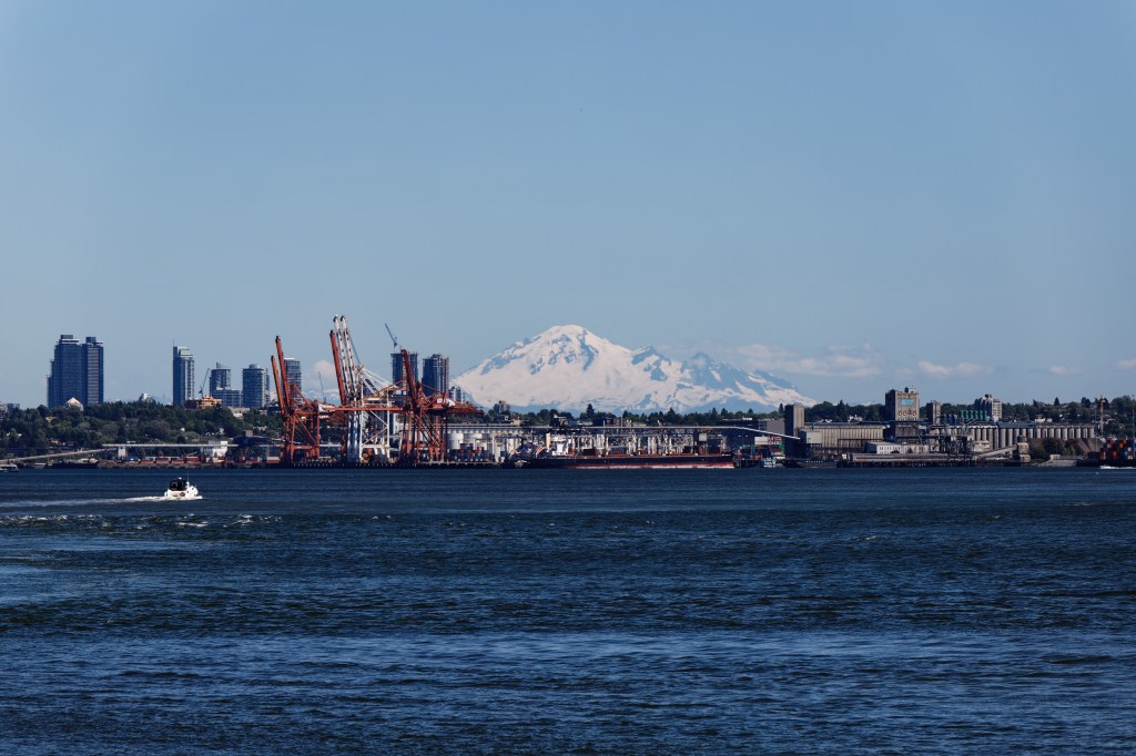 Blick auf den Hafen Vancouvers mit Kränen, Industrieanlagen und Bergen im Hintergrund. Im Vordergrund links fährt ein Motorboot.