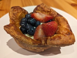 Ein Gebäck aus Blätterteig mit Vanillepudding, Blaubeeren und Erdbeeren liegt auf einem weißen Teller.