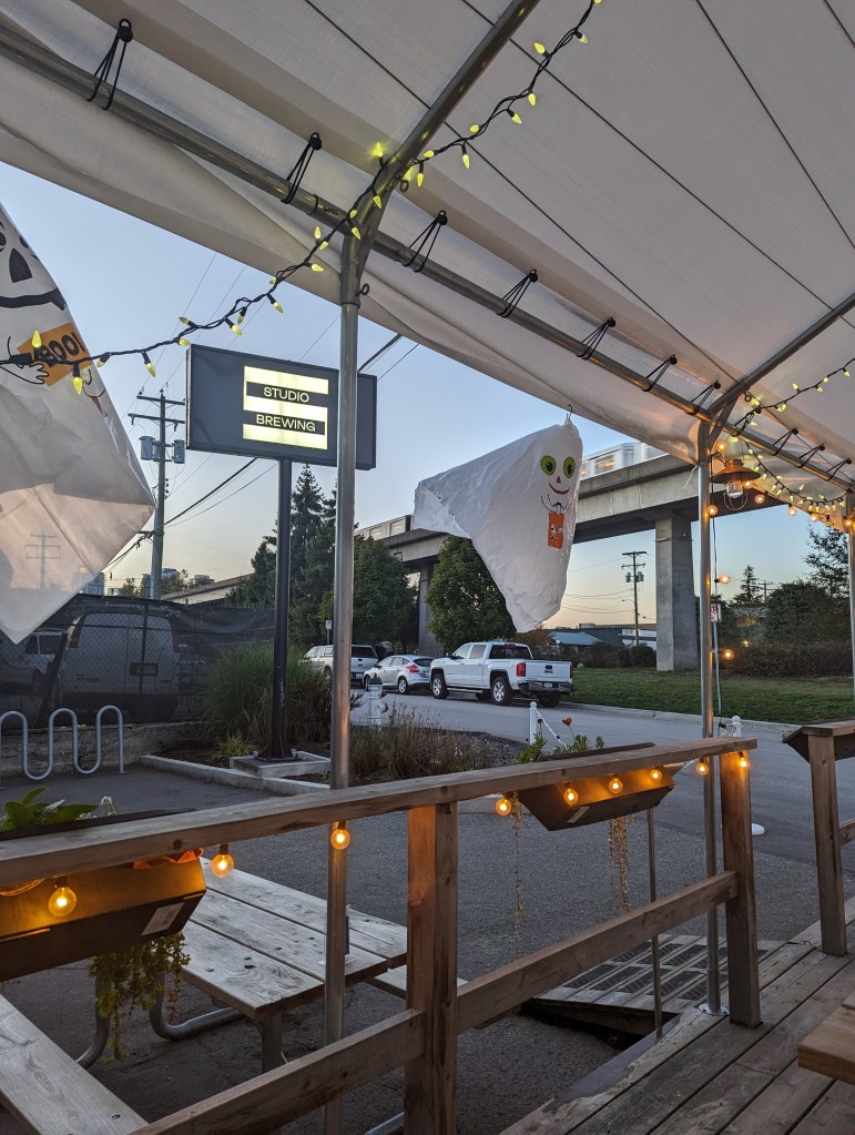 Der Patio von Studio Brewing in der Abenddämmerung: Die Lichterketten und das Schild der Brewery leuchten, Halloween-Plastik-Gespenster hängen vom Zeltdach. Im Hintergrund sieht man die Betonpfeiler und Gleise des Skytrains.