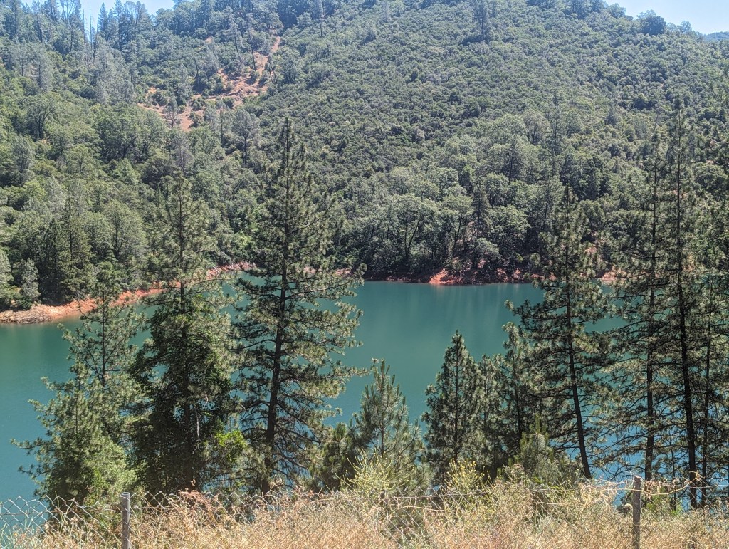 Im Vordergrund sieht man einen Zaun vor ein paar Bäumen. Im Tal darunter liegt ein grüner See, auf der anderen Uferseite erhebt sich ein kleiner Berg mit viel grün, Büschen und Bäumen.