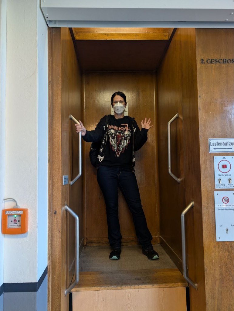 Eine Frau fährt in einem Paternoster-Aufzug und winkt dem Fotografen zu.