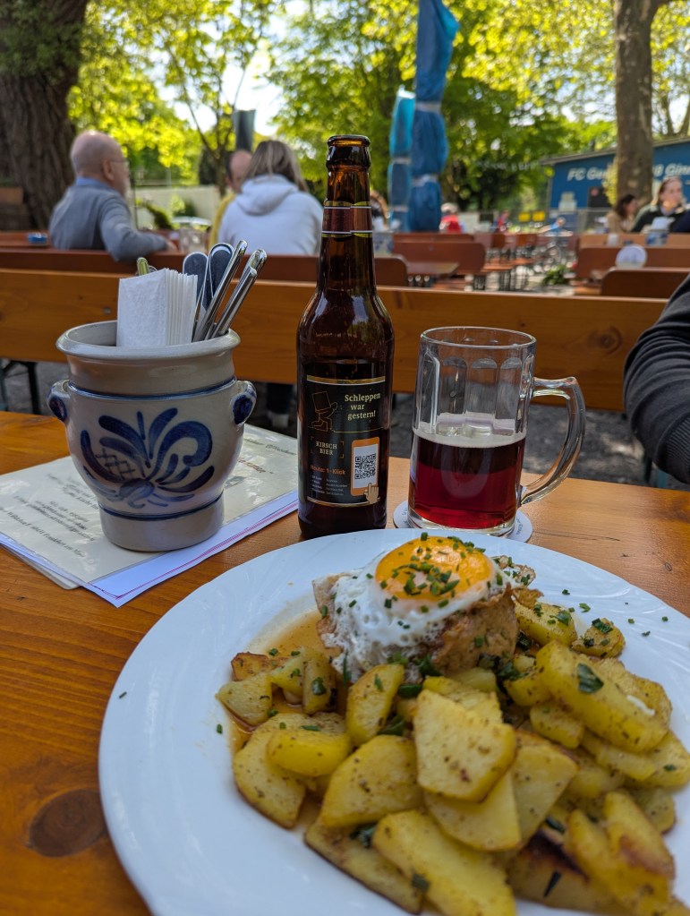 In einem Biergarten sieht man verschiedene Tische und Bänke. Auf dem vordersten Tisch stehen ein Gericht mit Spiegelei und Kartoffeln, dahinter ein Glas und eine Flasche Kirschbier. Daneben ein Bembel mit Geschirr und Servietten.