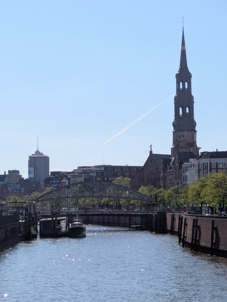 Ein Fluss, in dessen Wasser die Sonne glitzert. Am Ufer sieht man in der Ferne rechts Häuser und eine Kirche. LInks ist ein Boot festgemacht.