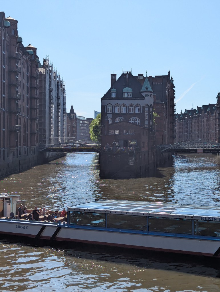Ein Boot wendet im Vordergrund auf einem Fluss vor einer kleinen Insel, auf der Häuser stehen. Rechts und links stehen ebenfalls Backsteingebäude. Die Ufer sind durch Brücken verbunden.