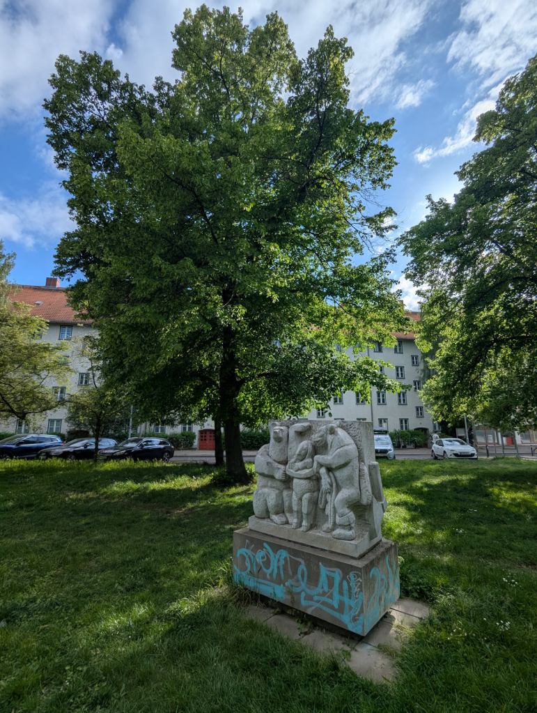 Grüne Wiese mit einer grauen Statue mit Sockel im Vordergrund. Darauf sind wohl Bären und ein Mädchen abgebildet. Dahinter steht ein hoher Baum und ein vierstöckiger Wohnblock mit roter Haustür und rotem Ziegeldach.