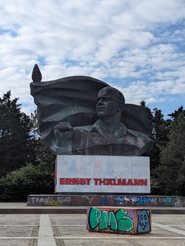Kollosalbüste von Ernst Thälmann, dem ehemaligen Vorsitzenden der Kommunistischen Partei Deutschlands (KPD), der von den Nazis ermordet wurde. Die Büste steht im Bezirk Prenzlauer Berg, im Osten Berlins. Thälmann hat die rechte Hand zur Faust geballt und blickt nach rechts mit ernstem Blick, etwas gerunzelter Stirn. Der Sockel des Denkmals ist mit Graffiti überzogen. Davor steht ein quaderförmiges Betonobjekt, in dem die Worte "Kopf Faust Fahne" eingraviert sind. Auch dieses Objekt ist mit bunter Graffiti überzogen.
