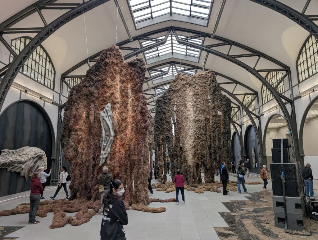 Die Installation "Embrace" der tschechischen Künstlerin Klára Hosnedlová. Man sieht von der Decke einer großen Halle hängenden Flachs. Auf diesen miteinander verknüpften Flachsteilen sind teilweise Objekte angebracht. Ähnliche Objekte, die wie Fossilien von Dinosauriern anmuten, hängen an den Wänden.