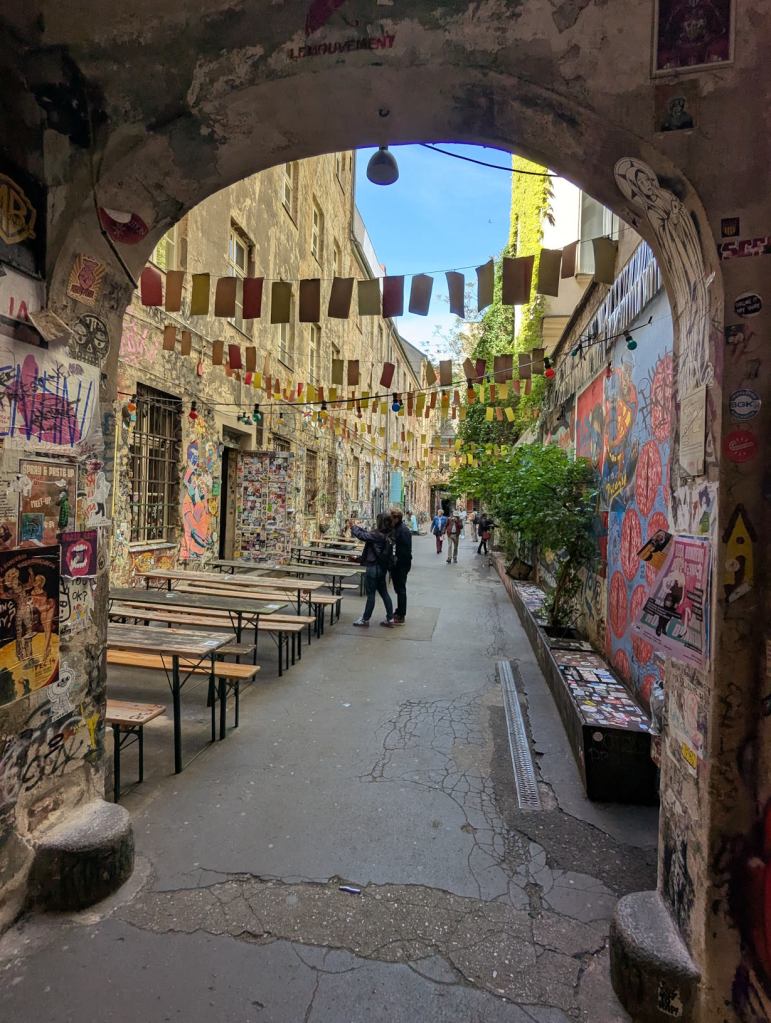 Eingang zum Innenhof von Haus Schwarzenberg. Die Wände sind mit vielen Postern beklebt. Links im Hof stehen Bierbänke und -tische. Über den Platz sind Leinen mit bunten Zetteln gespannt. Rechts wurden um junge Bäume dunkle Holzbänke gebaut.