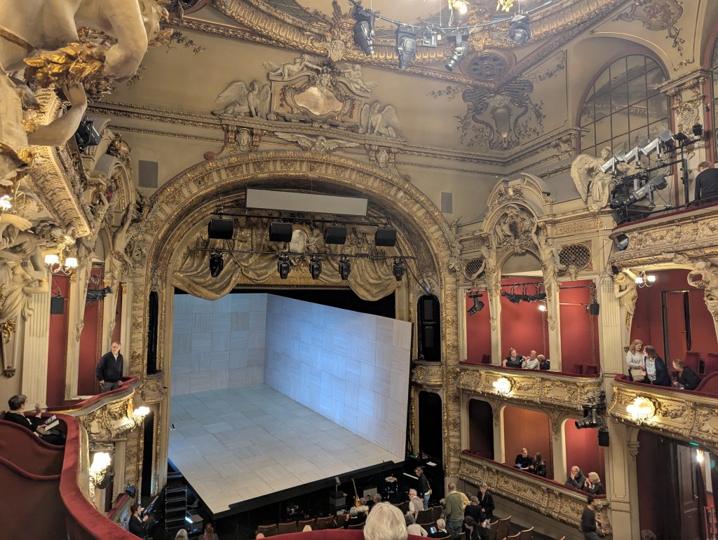 Man sieht einen üppig ausgestatteten Theatersaal mit goldenem, verschnökeltem Dekor, rotem Samt an den Wänden und an den Brüstungen der Logen sowie einem goldenem Vorhang, der hochgezogen ist. Er gibt den Blick auf eine komplett leere Bühne frei.