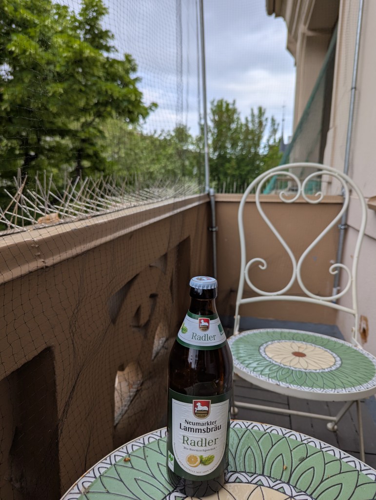 Auf einem Balkon steht eine Flasche Neumarkter Lammsbräu Radler auf einem Gartentisch. Dahinter steht ein Gartenstuhl. Der Balkon ist von einem Netz umspannt, das Vögel davon abhalten soll, auf den Balkon zu kommen. Links neben dem Balkon sieht man einige Baumwimpfel.