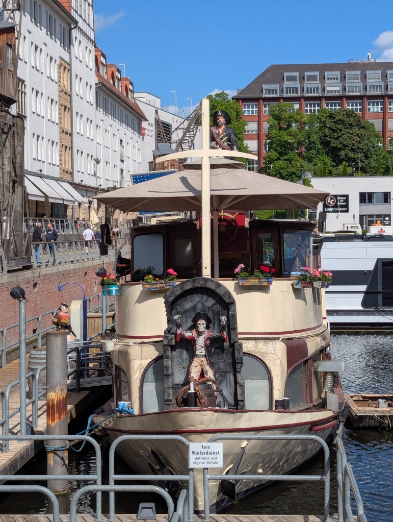 In einem Hafen liegt ein kleiner, eierschalenfarbener Ausflugsdampfer, der über zwei Piratenfiguren verfügt. Die eine ist am Bug des Schiffes angekettet, besteht aus einem Skelett, trägt einen schwarzem Hut und die gängige Piratenkleidung. Die andere ragt vom Heck des Schiffes über das Dach vor: Der Pirat hat ein bärtiges Gesicht, eine Augenklappe und ebenfalls einen schwarzen Hut. Außerdem hat er an der rechten Hand einen silbernen Haken.