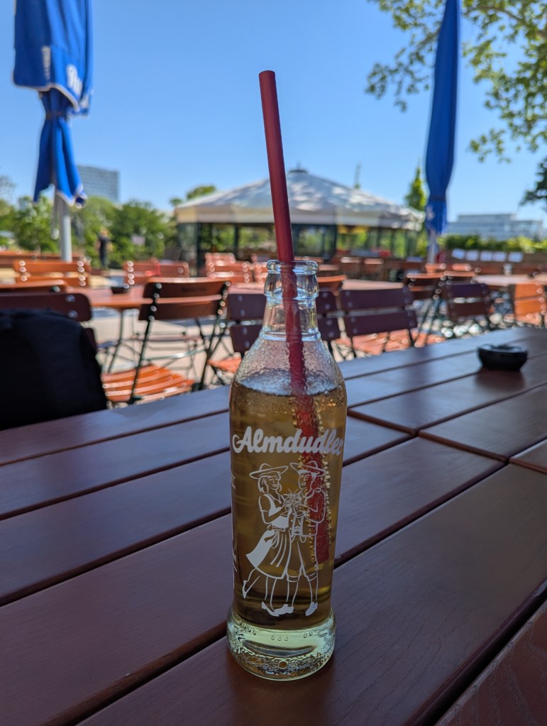 Eine Flasche Almdudler-Limonade steht auf einem Tisch in einem Biergarten. Dahinter sind Stühle, weitere Tische, Sonnenschirme und Bäume zu sehen.