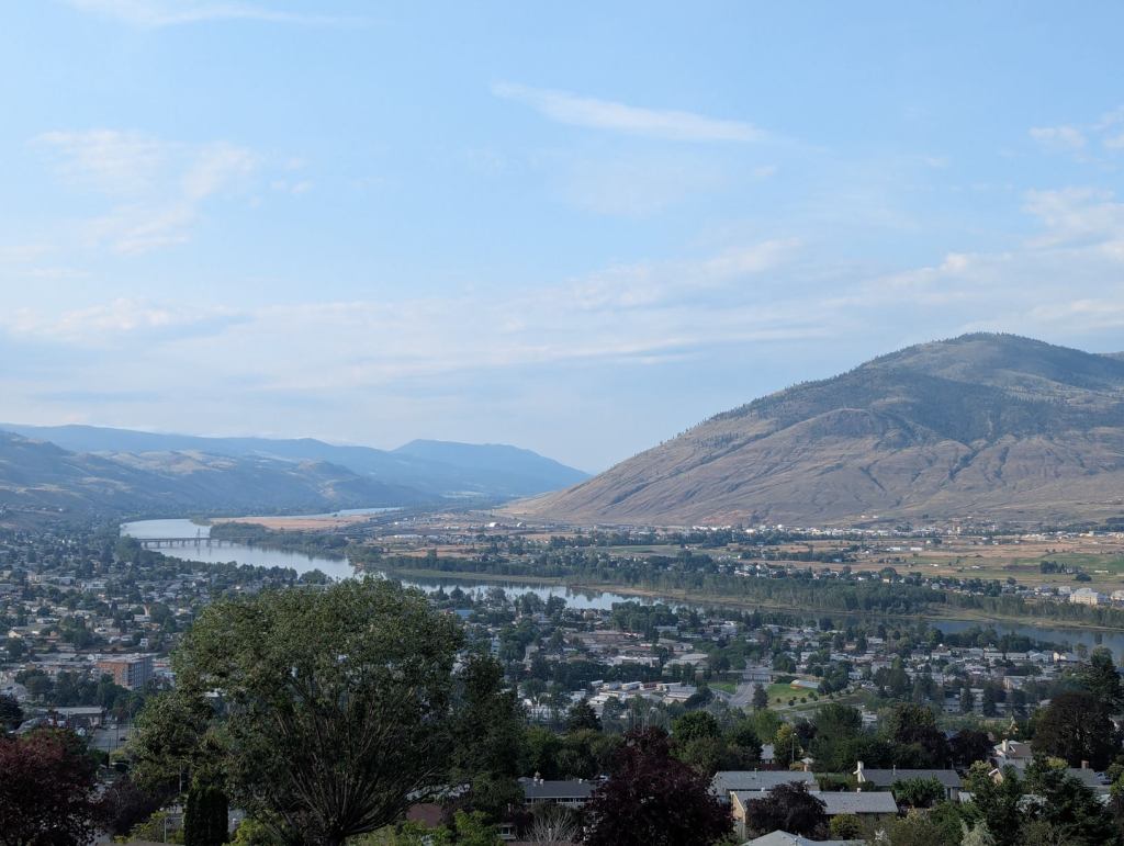 Ein Tal mit Bergen, die mit Bäumen und Sträuchern bewachsenen sind. In der Mitte schlängelt sich ein Fluss durch das Tal. einem An dessen Ufern stehen Häuser. Im Himmel gibt es einige Schleierwolken und etwas Sonnenlicht zu sehen.