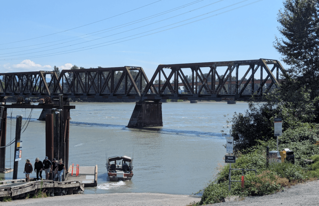 Eine alte eiserne Brücke, über die wohl kein Zug mehr fährt, führt über einen Fluss. Im Vordergrund kann man ein Boot sehehn, das gerade eine Gruppe von Menschen an Land gebracht hat.