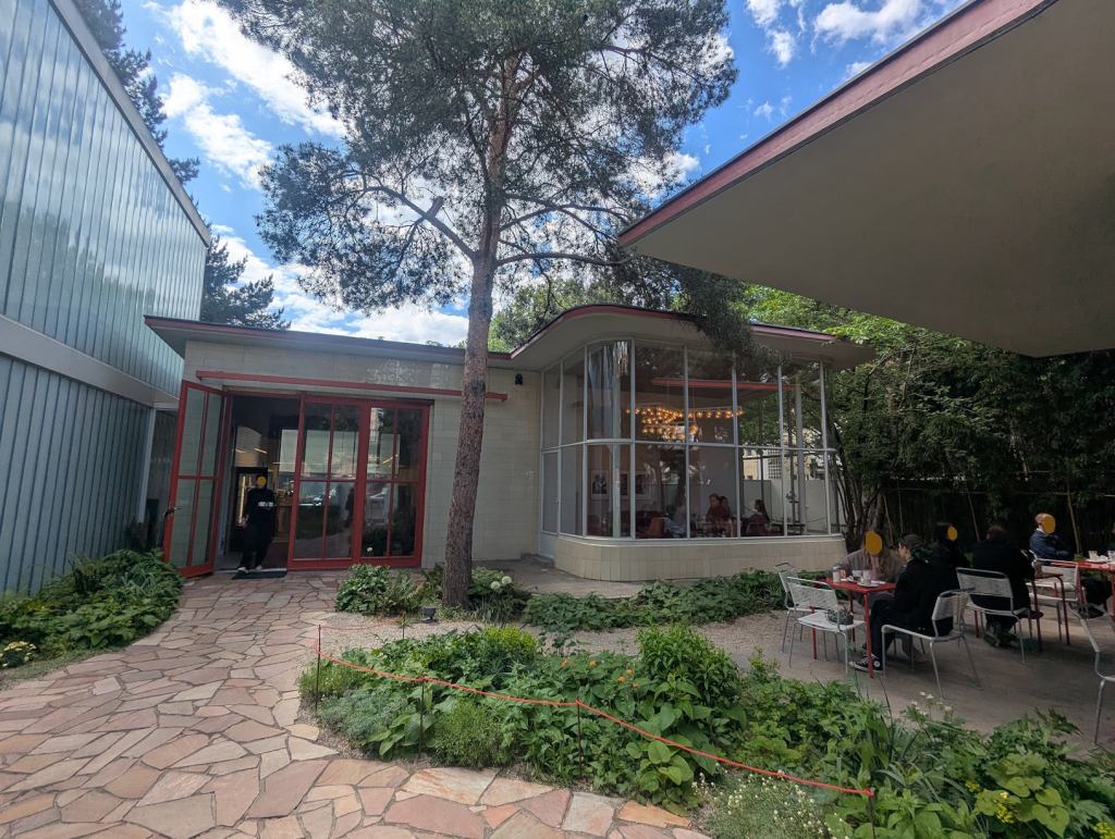 Eine Tankstelle aus den 50ern Jahren, die jetzt als Café genuetzt wird. Vor dem Gebäude sind Steinplatten verlegt. Der Bau ist weiß gestrichen und hat rote Türrahmen.