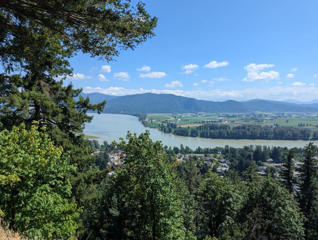 Aussicht von einer Anhöhe auf ein Tal mit Fluss, grünen Bergen, Wiesen und Bäumen.