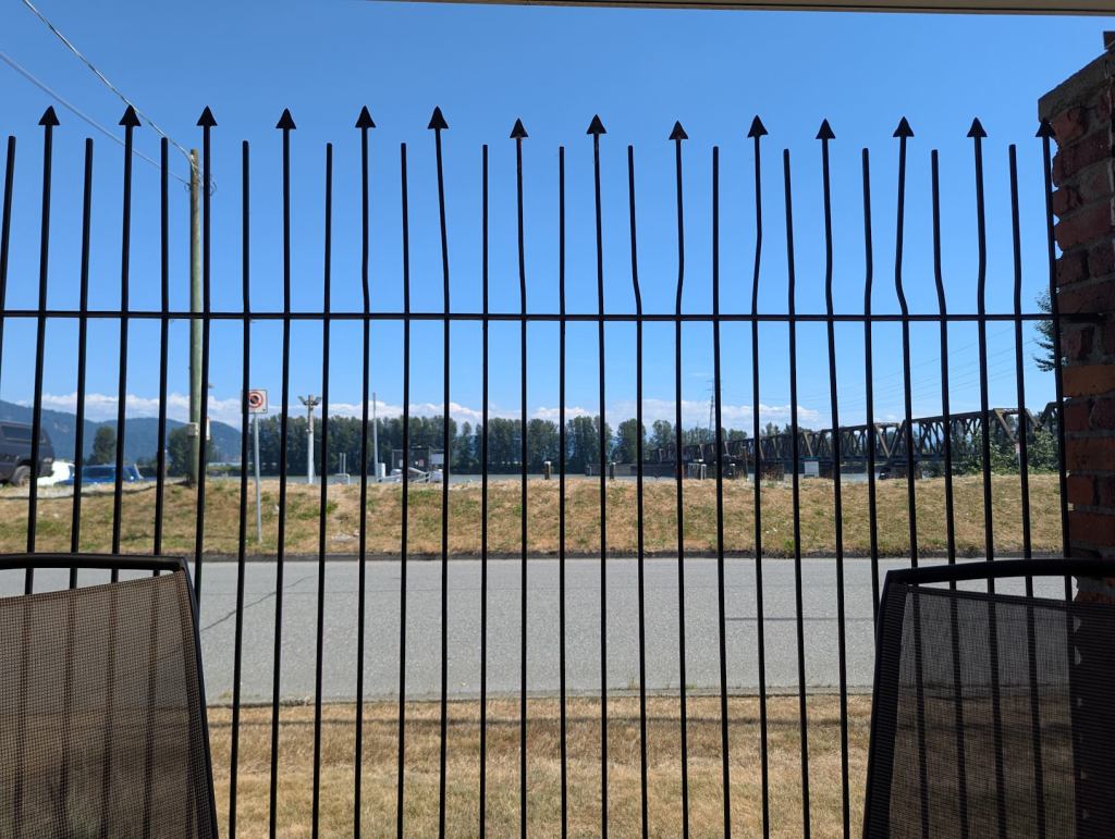 Durch einen Zaun kann man eine Straße und das dahinter liegende Flussufer sehen. Seitlich der Straße gibt es zwei Streifen von Gras, die sehr trocken und daher gelbfarben sind.