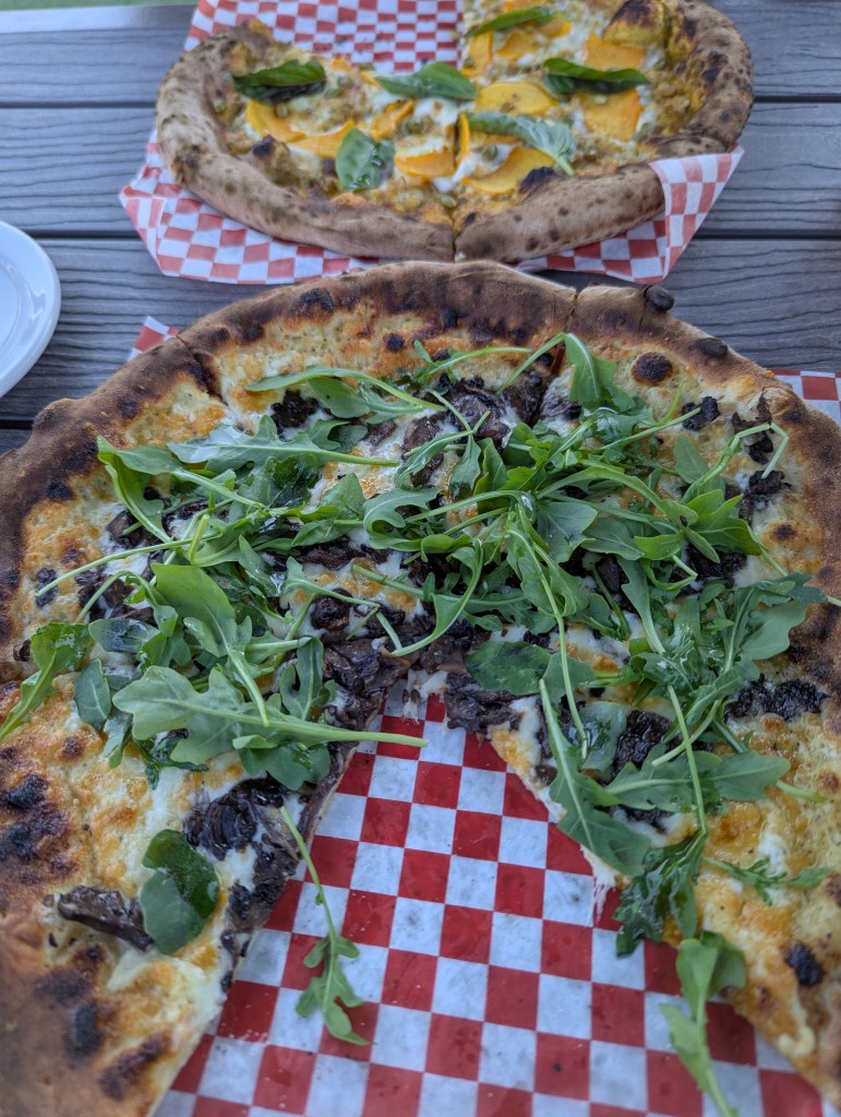 Im Vordergrund eine Pizza mit Wildpilzen und Rucola, im Hintergrund eine Pizza mit Kürbis und Blättern, die wie Basilikum aussehen. An beiden Pizzen fehlt jeweils ein Stück. 