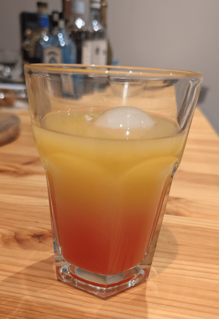 Ein Glas mit Tequila Sunrise inklusive Eiswürfel steht auf einem Holztisch.