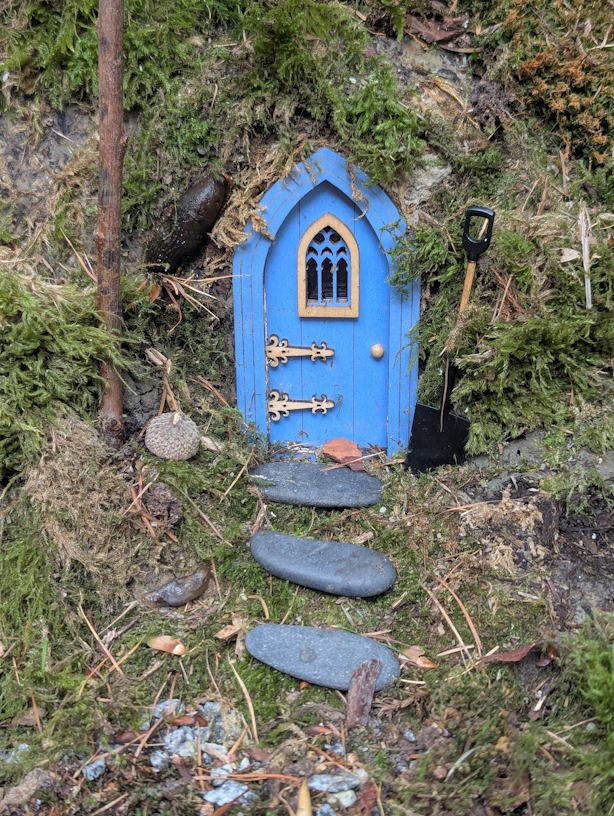 In einem Baumstamm wurde eine blaue kleine Tür eingelassen. Daneben steht eine Schaufel und drei Steine markieren den Weg zur Tür.