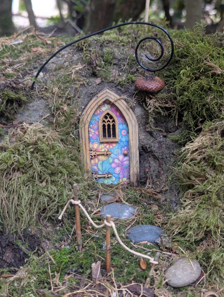In die Wurzeln bzw. das Erdreich unterhalb eines Baumes hat jemand eine kleine Tür - wie für Zwerge - eingebaut. Diese besteht aus einem hellbraunen giebelförmigen Türrahmen. Dazu ist ein ebenso giebelartiges Fenster in die Mitte der Tür eingelassen, das mit Sprossen versehen wurde. Zwei kunstvoll verzierte Schlösser ragen von der rechten Seite bis in die Mitte der Tür. Auf der linken Seite kann man einen Türknauf sehen. Die Tür selbst ist mit Blumen in blauen Farben, hellem Lila und gelb bemalt. Darunter markieren Steine den Pfad zur Tür. Rechts ist zudem ein Geländer aus Holzpfosten, die mit einem Seil verbunden sind, angebracht. Über der Tür ist ein wie Metall aussehenden Lampenhalter befestigt, der in eine Spirale mündet und über die Tür ragt. Daran hängt nicht wie erwartet eine Lampe, sondern der braune Fruchtbecher einer Eichel.