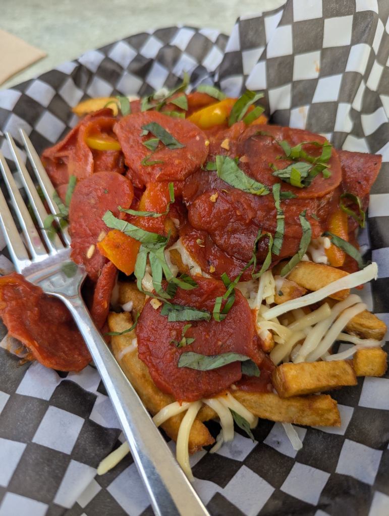 Ein Korb, der mit einem Papier mit Schachbrettmuster, ausgefüllt ist. Darauf liegen Pommes Frites, darüber wurde eine Tomatensauce gegossen. Wurstscheiben, ein paar Paprikawürfel und Streukäse sieht man ebenfalls.
