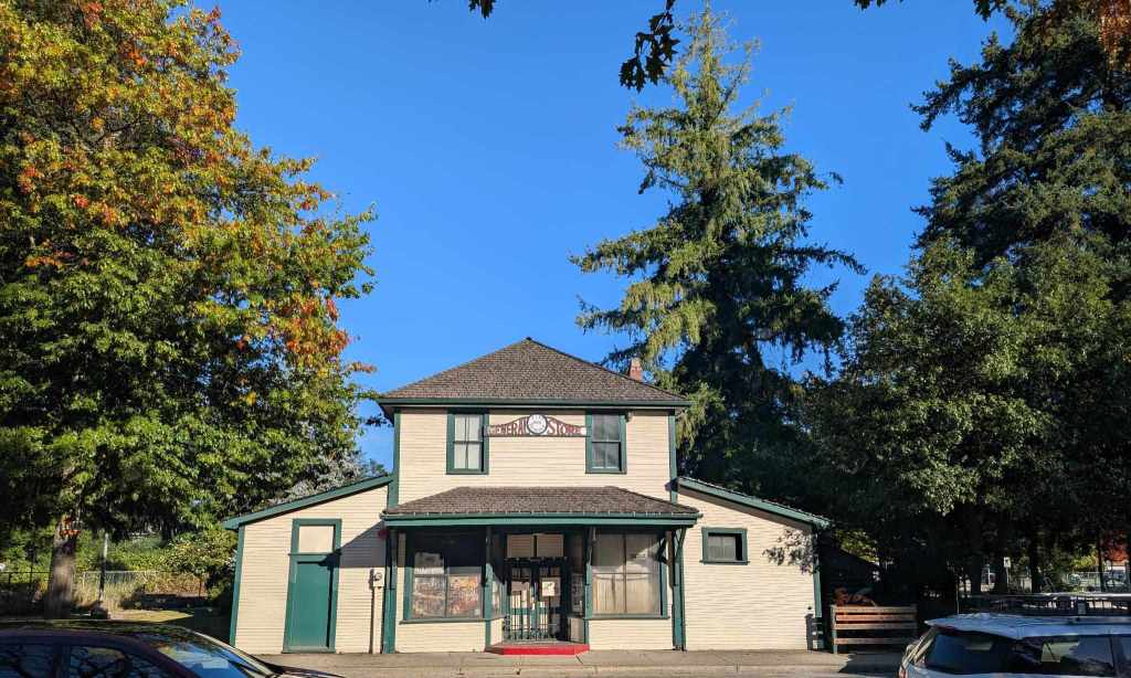 Ein zweigeschossiges hellbraunes Holzhaus, auf dem "General Store" sowie "Pitt Meadows 1886" geschrieben steht. Eine weitere Aufschrift "Museum" kann man erkennen.