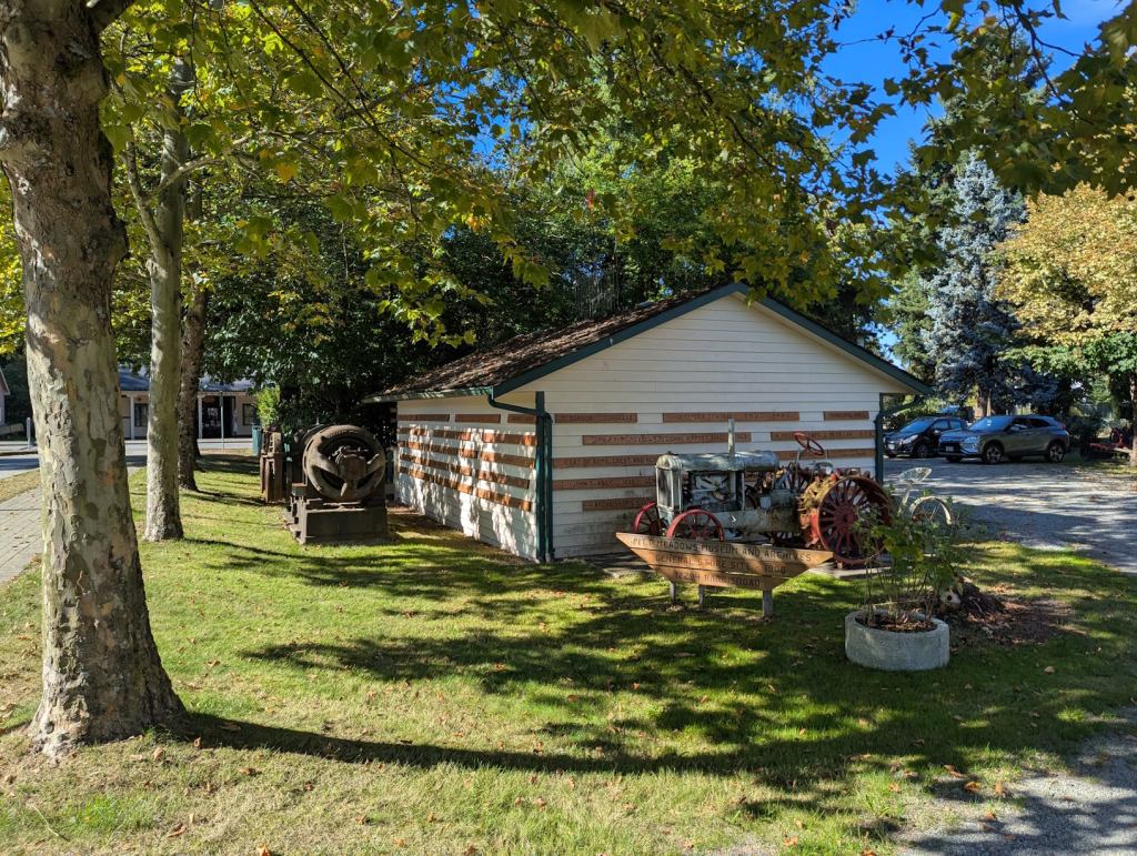 Auf einer Rasenfläche mit Bäumen steht ein kleines weißes Holzgebäude, davor ein Schild mit der Aufschrift "Pitt Meadows Museum and Archives". Davor steht ein alter Traktor.