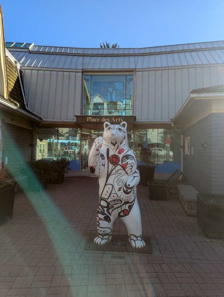 Die Statue eine weißen Bären steht vor eine Art Community Centre, das den Namen "Place des Arts" trägt. Der Bär trägt schwarz-rote Bemalungen nach typisch indigenen Mustern der kanadischen First Nations. Die Statue eine weißen Bären steht vor eine Art Community Centre, das den Namen "Place des Arts" trägt. Der Bär trägt schwarz-rote Bemalungen nach typisch indigenen Mustern der kanadischen First Nations.
