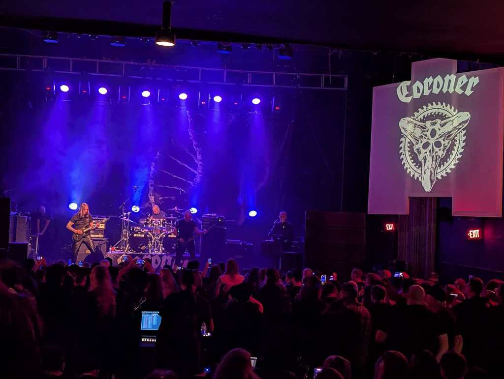 Die Band Coroner spielt auf der Bühne. Daneben ist das Logo der Band an die Wand projiziert. Der Zuschauerraum ist gut gefüllt.