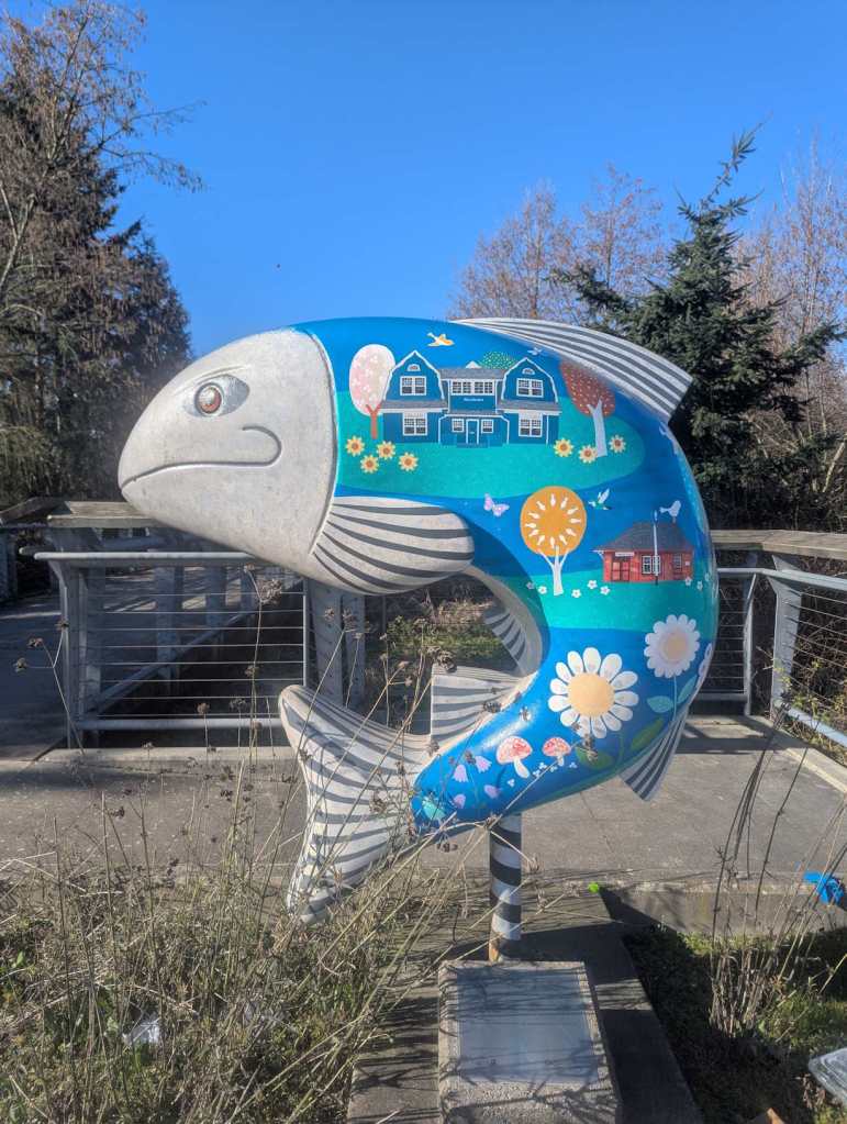 Eine Fischstatue am Eingang eines Parks, die mit Häusern, Bäumen und Blumen bemalt wurde. Der Fisch hat einen fröhlichen Gesichtsausdruck. Eine Fischstatue am Eingang eines Parks, die mit Häusern, Bäumen und Blumen bemalt wurde. Der Fisch hat einen fröhlichen Gesichtsausdruck.