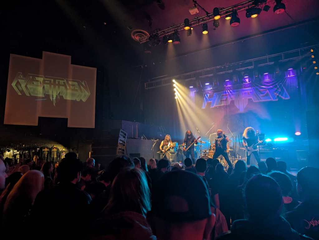 Die Band Heathen spielt auf der Bühne. Viele Zuschauerinnen und Zuschauer stehen davor.