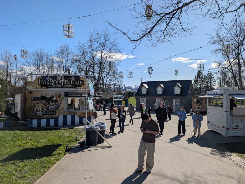 Ein Park, in dem verschiedene Food Trucks stehen und Besucherinnen und Besucher herumlaufen. Auf der linken Seite sieht man eine Wiese und einen Wagen mit der Aufschrift "La Poutinerie", auf dem man große gemalte Körbchen mit Pommes sehen kann.