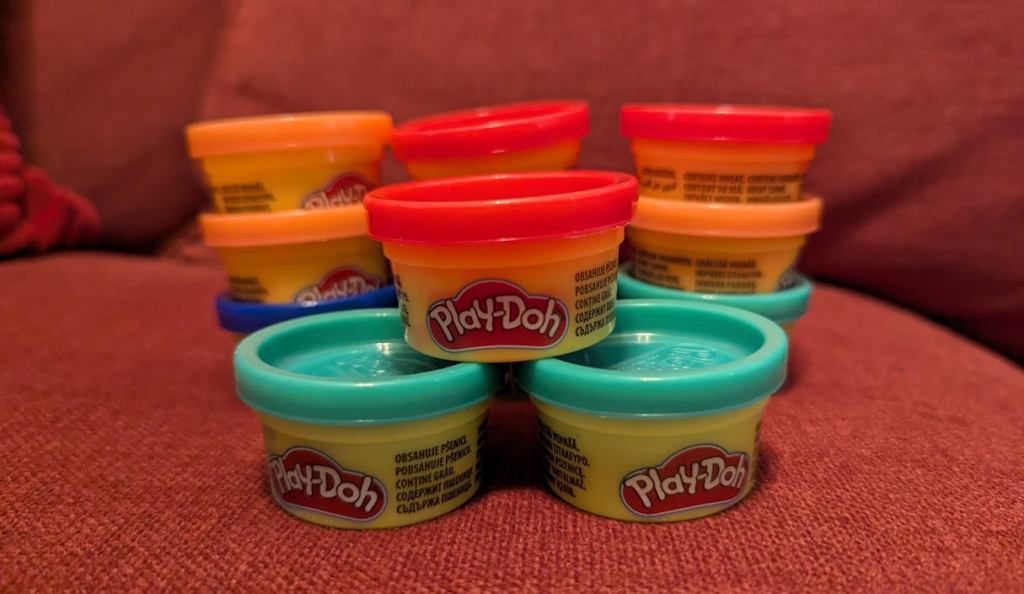 Verschiedene bunte Plastikeimerchen mit Kinder-Knetmasse stehen aufgestapelt auf einer Couch. Die kleinen Eimer haben grüne, rote und orange Deckel. Sie tragen die Aufschrift "Play-Doh". Verschiedene bunte Plastikeimerchen mit Kinder-Knetmasse stehen aufgestapelt auf einer Couch. Die kleinen Eimer haben grüne, rote und orange Deckel. Sie tragen die Aufschrift "Play-Doh".