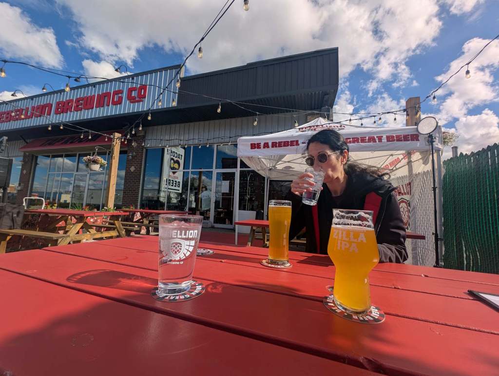 Eine Frau sitzt an einem Tisch und trinkt aus einem Glas mit Wasser. Daneben stehen Gläster mit Bier. Im Hintergrund sieht man einen Sonnenschirm mit der Aufschrift "Be a rebel".