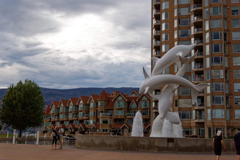Im Vordergrund ist ein Brunnen mit verschiedenen Wasserfontänen und eine Skulptur mit drei Delfinen, die ineinander springen, zu sehen. Dahinter zwei Wohnhochhäuser rechts und links eine Wohnzeile mit vielen verschiedenen Wohnungen.