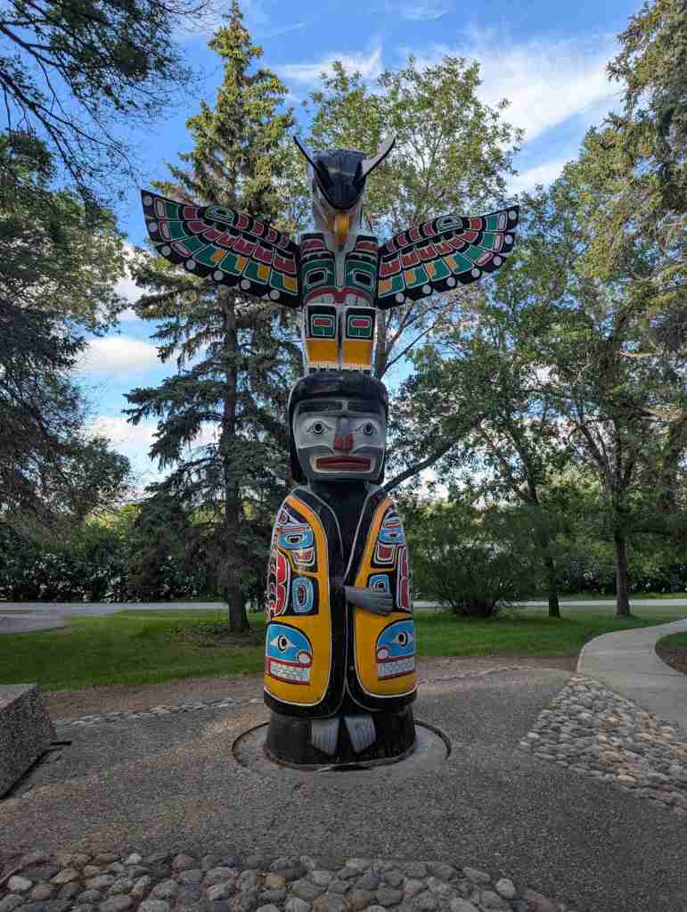 Ein bunter Totem-Pfahl in den Farben orange, blau, schwarz, grün und weiß in einem Park.