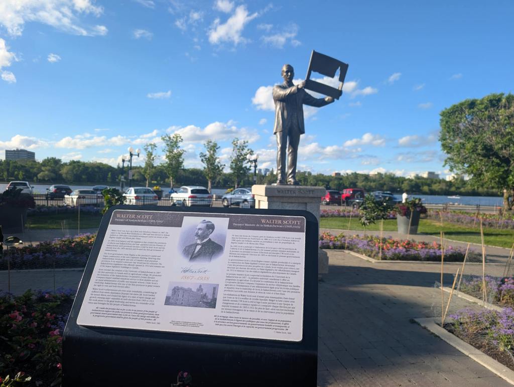 Die Statue eines Mannes steht auf einem Sockel, sie hält die Umrisse des Legislativ-Gebäudes in der Hand. Die Statue ist diesem Gebäude zugewandt. Sie ist in einen Park eingebettet. Hinter der Statue ist ein See.