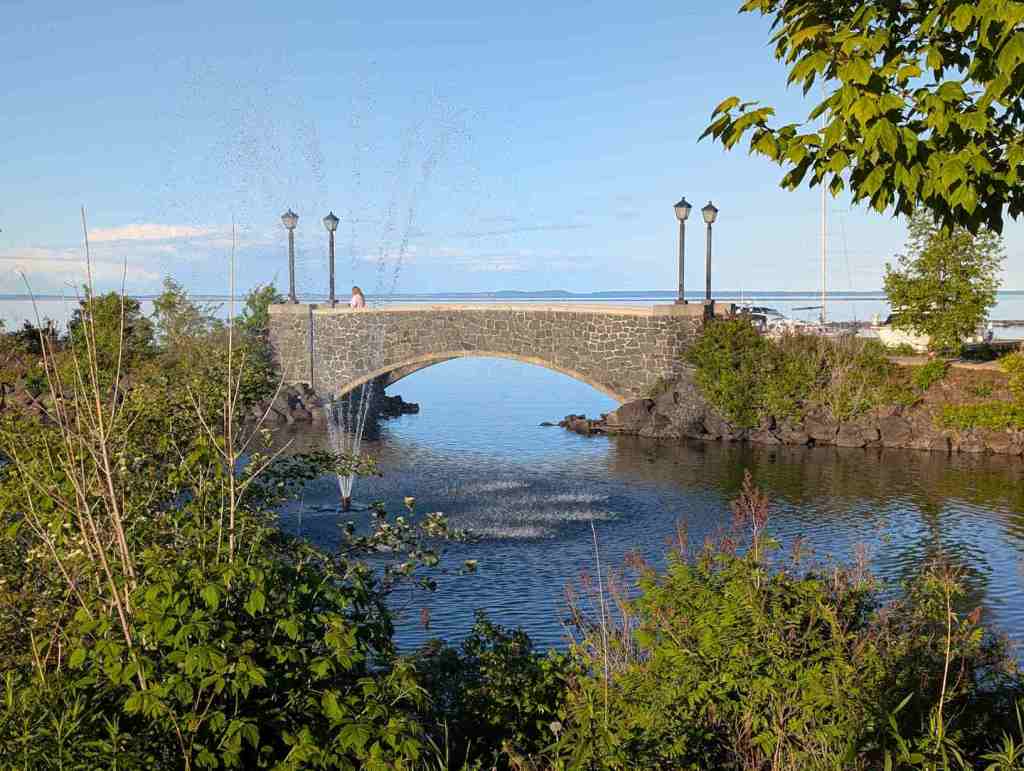 Eine kleine steinerne Brücke über den Arm eines Sees. In der Bucht sprüht eine Fontäne Wasser nach oben.