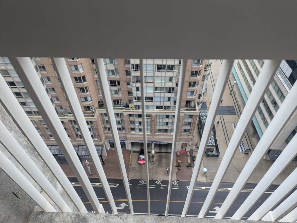 Die Gitter eines Balkons. Es geht tief hinunter auf eine Straße. Auf der anderen Seite steht eine Wohnhochhaus mit vielen Fenstern und Balkonen.