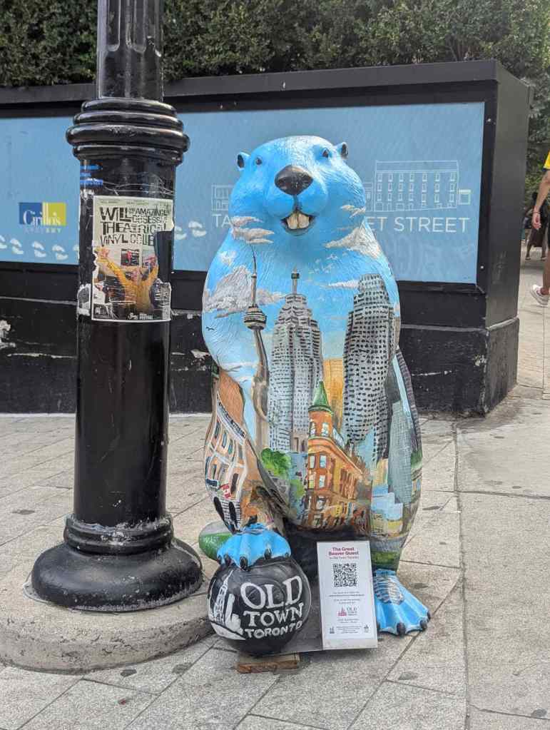 Eine blaue Biberfigur steht an einer Straßenlaterne. Der Biber hat die Skyline Torontos auf den Körpfer gemalt. Er hat den Fuß auf einem Ball platziert, der den Schriftzug "Old Town Toronto" trägt.