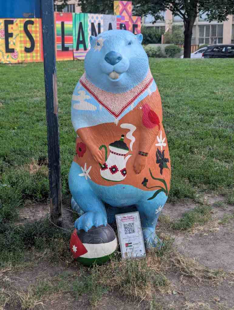 Eine blaue Biberfigur, die eine orangefarbene Jacke trägt und eine Kaffeekanne mit dampfenden Kaffee in der Pfote hält. Der Biber hat den Fuß auf einer Kugel mit der jordanischen Flagge gestellt.
