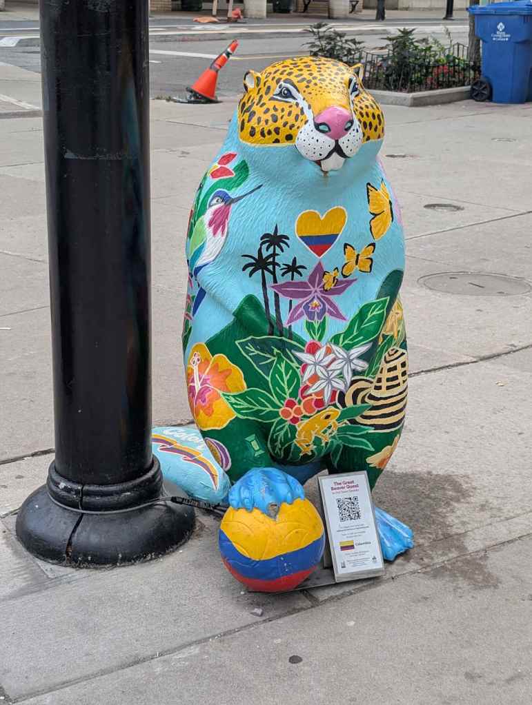 Eine bunte Biberfigur mit dem Kopfmuster eines Leoparden. Vögel, Schmetterlinge und Blumen zieren den Körper. Der Fuß steht auf einer Kugel mit der kolumbianischen Flagge.