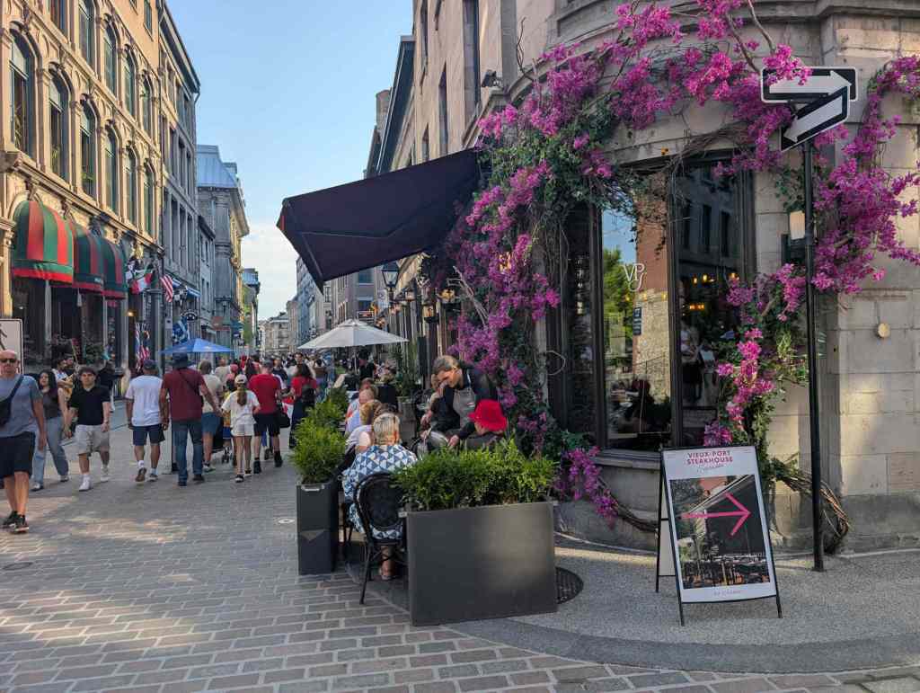 Ein mit pinkfarbenen Blumenranken verziertes längliches Fenster eines Steakhauses. Daneben sitzen Restaurantbesucher*innen an Tischen in einem Gässchen.