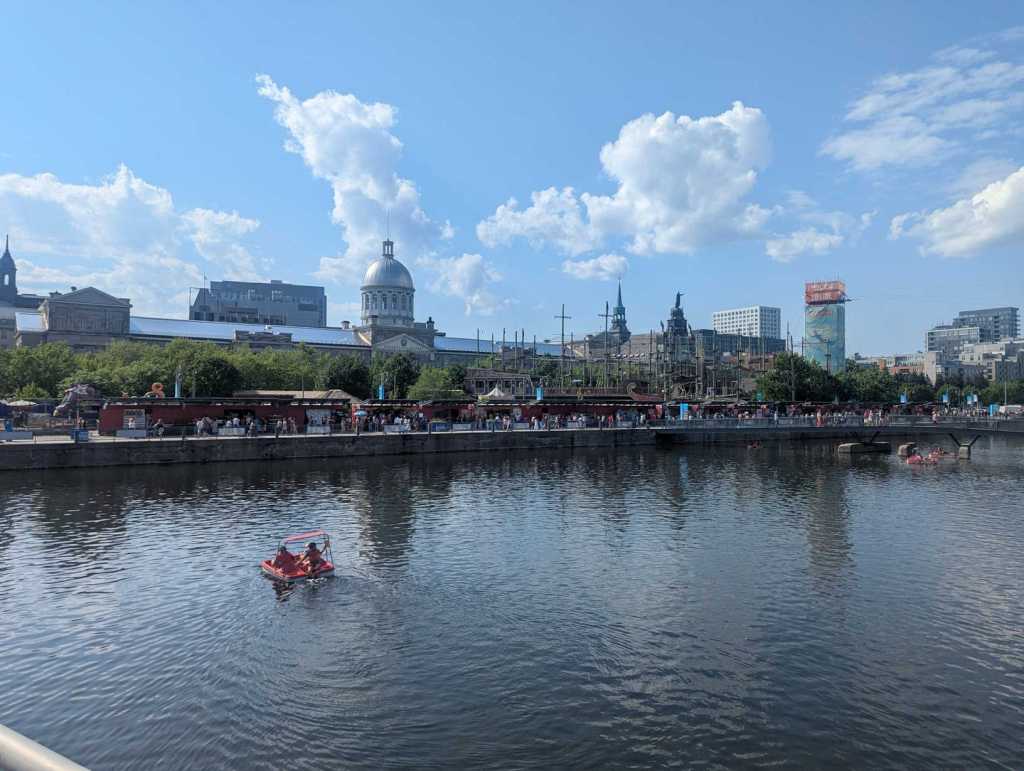 Ansicht eines Hafengeländes mit vielen Buden vom Wasser aus. Im Hintergrund sieht man historische Gebäude, unter anderem mit einer Kuppel.