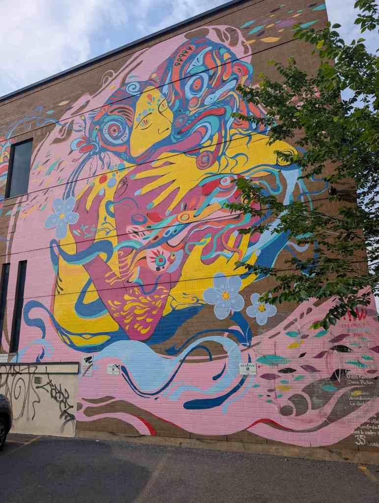 Buntes Mural einer Frau, die wohl ein Kind in den Armen hält. Alles verschmilzt mit Blumen und Fischen. Es erinnert an indigene Motive.