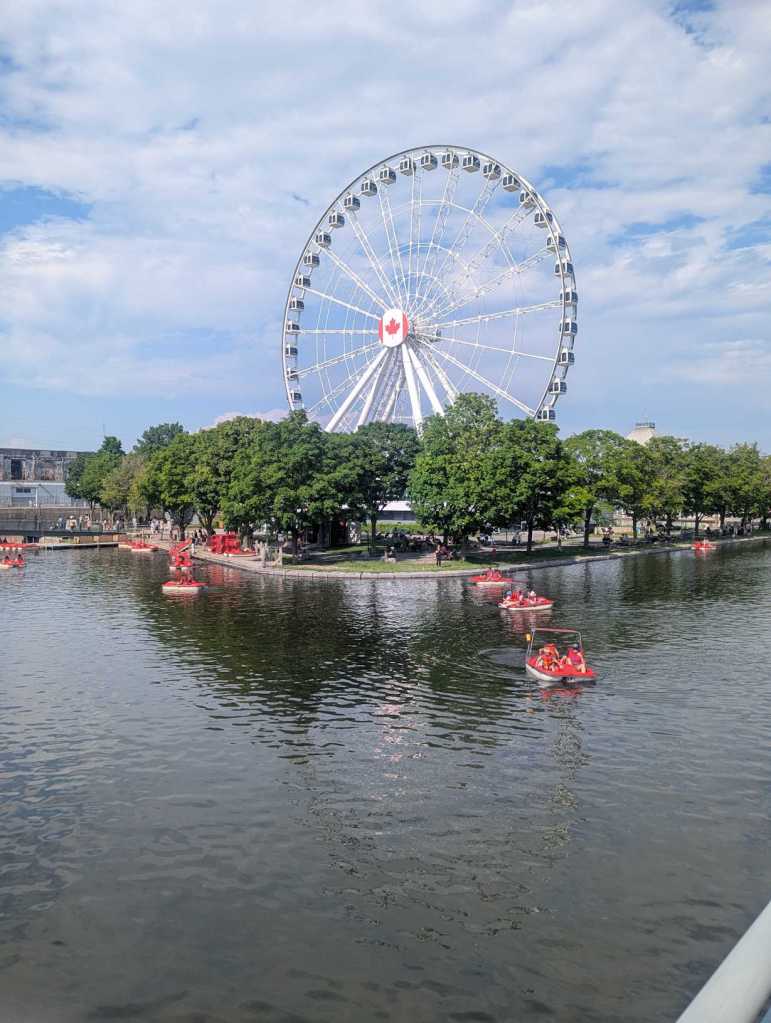 Riesenrad auf einem vorgelagerten Stück Land vor einem Hafen. Davor sind viele rote Tretbote zu sehen.