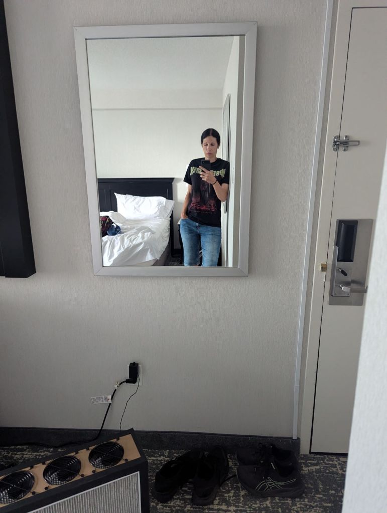 Selfie einer Frau im Hotelzimmer. Im Spiegel sieht man die Frau und Teile eines Betts. Ein Luftfilter steht auf einem Teppichboden.