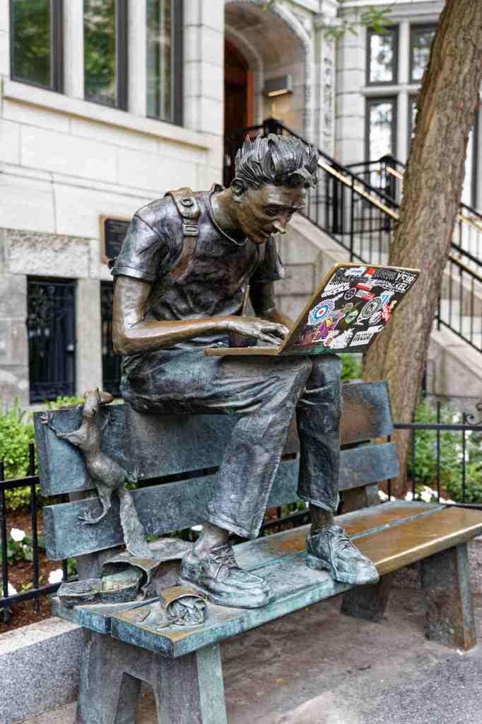 Skulptur im öffentlichen Raum: Ein junger Mann sitzt mit seinem Laptop auf einer Bank. Neben ihm eine Tasche, eine Schachtel Pommes und eine Ratte.
